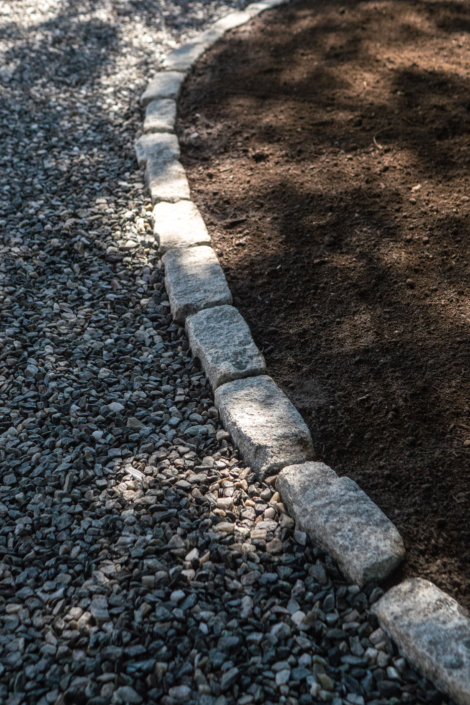 Curving cobblestone border separating crushed bluestone driveway material and fresh loam.