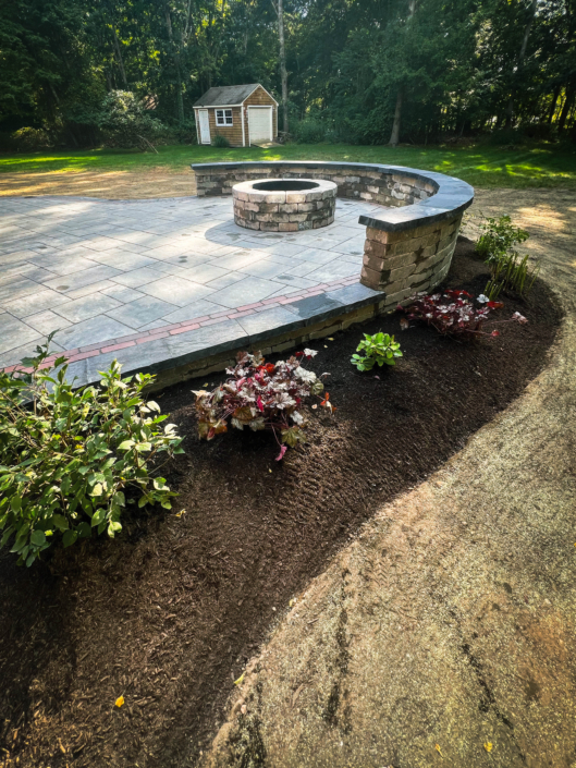 Raised rounded backyard patio with a seating wall along the far side, a circular fire pit within reach of the seating wall, and an ornamental garden wrapping around the patio.