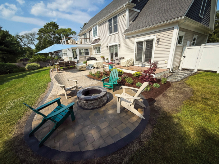 Circular patio extension featuring a stone fire pit and Adirondack chairs, with an ornamental garden between the patio and an upper deck to the home next to a bluestone path leading to an outdoor shower.
