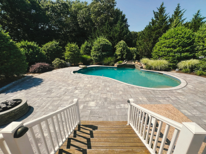 Point of view from the top of a flight of deck stairs leading to a paver patio surrounding an in-ground swimming pool with natural stone features.
